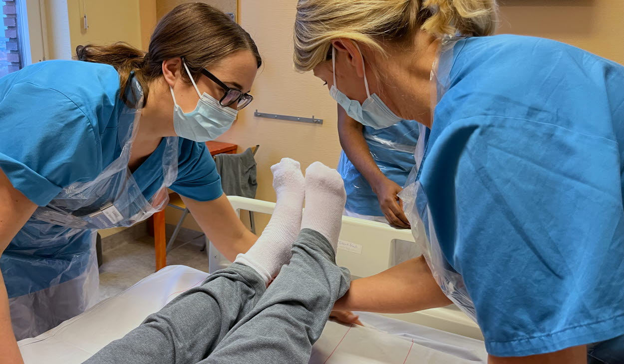 Två vårdpersonal med munskydd står böjd över fotändan i en säng, där det ligger en patient. De lyfter en fot på en patient, foten har vit socka på sig. Endast underbenen syns. 
