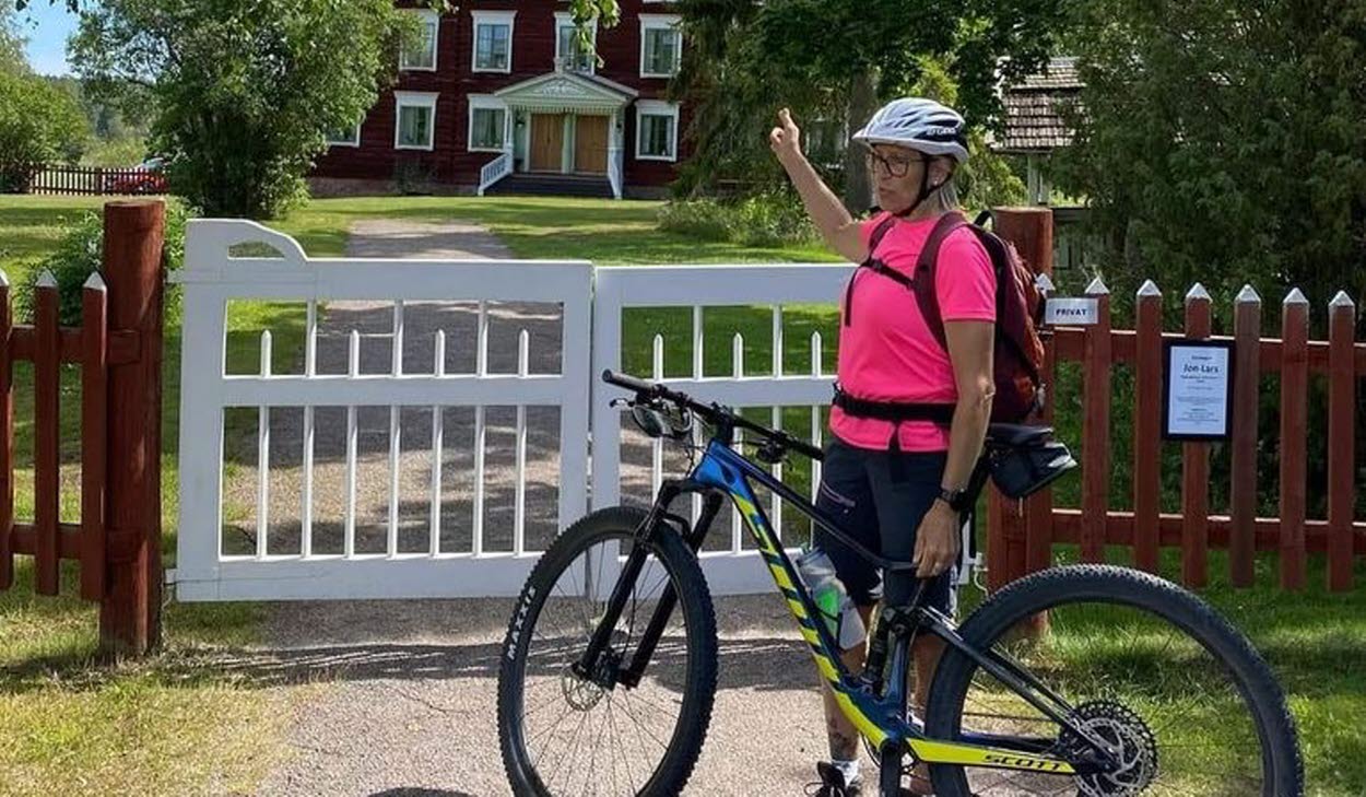 Person på cykel utanför en vit grind och rött staket. I bakgrunden är en Hälsingegård. 