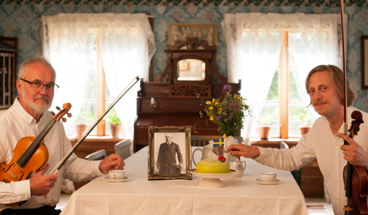 Två personer sitter vid ett bord med varsitt instrument och tittar in i kameran.