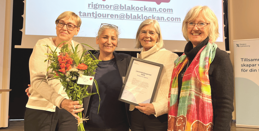 Fyra personer i halvfigur står framför en skärm med ordet Rigmor. De har blommor och diplom i händerna