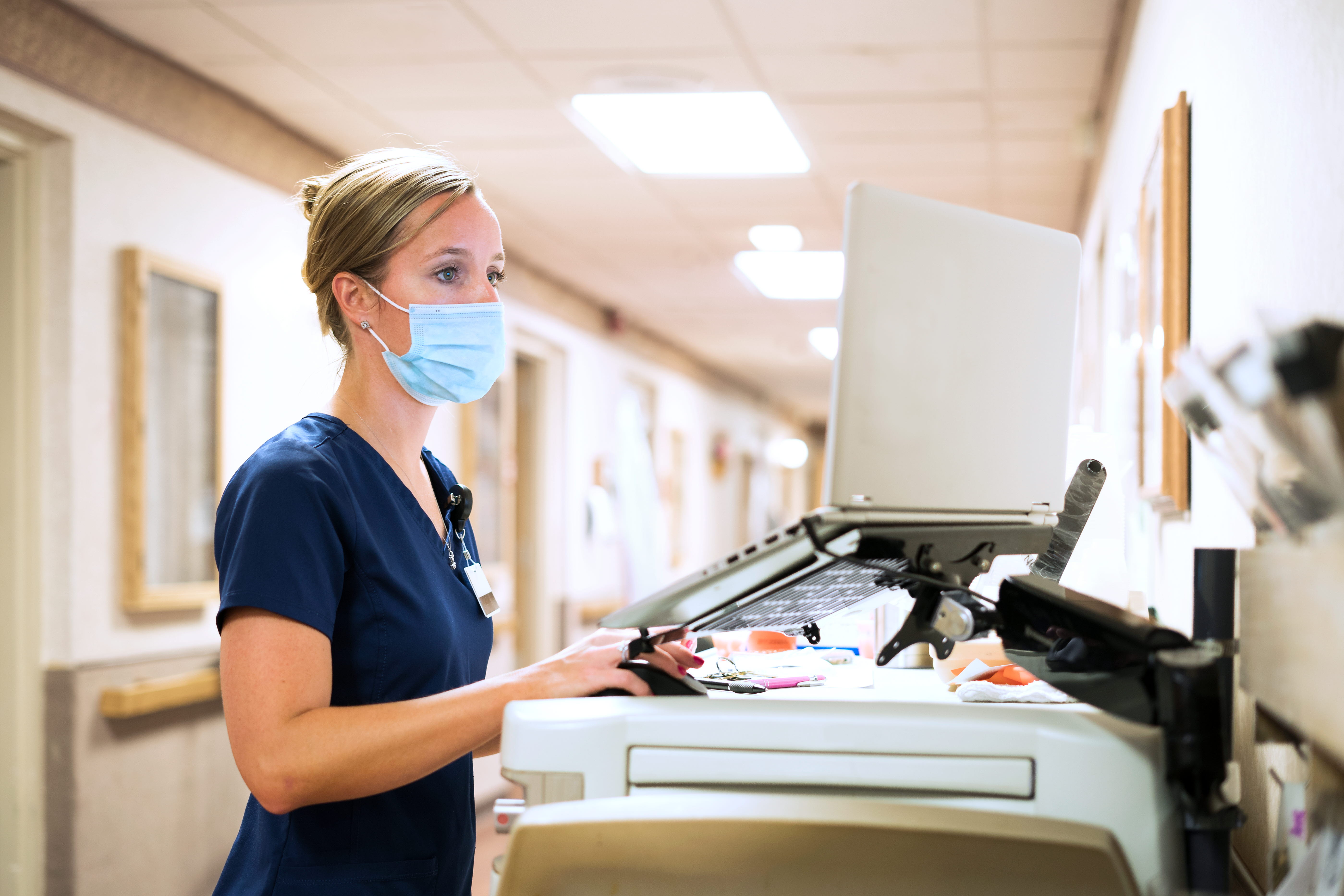 Registered nurse wearing a face mask documents medicine provided to a patient into a laptop computer at the nursing station at work, Indiana, USA