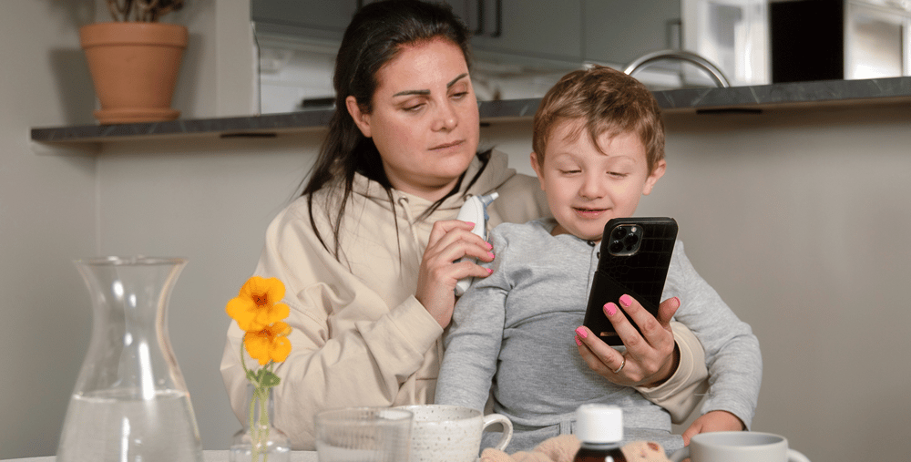 Mamma med barn i knä håller i telefon och termometer