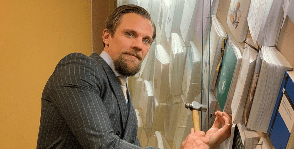 En man i kostym håller i en hammare framför en anslagstavla.