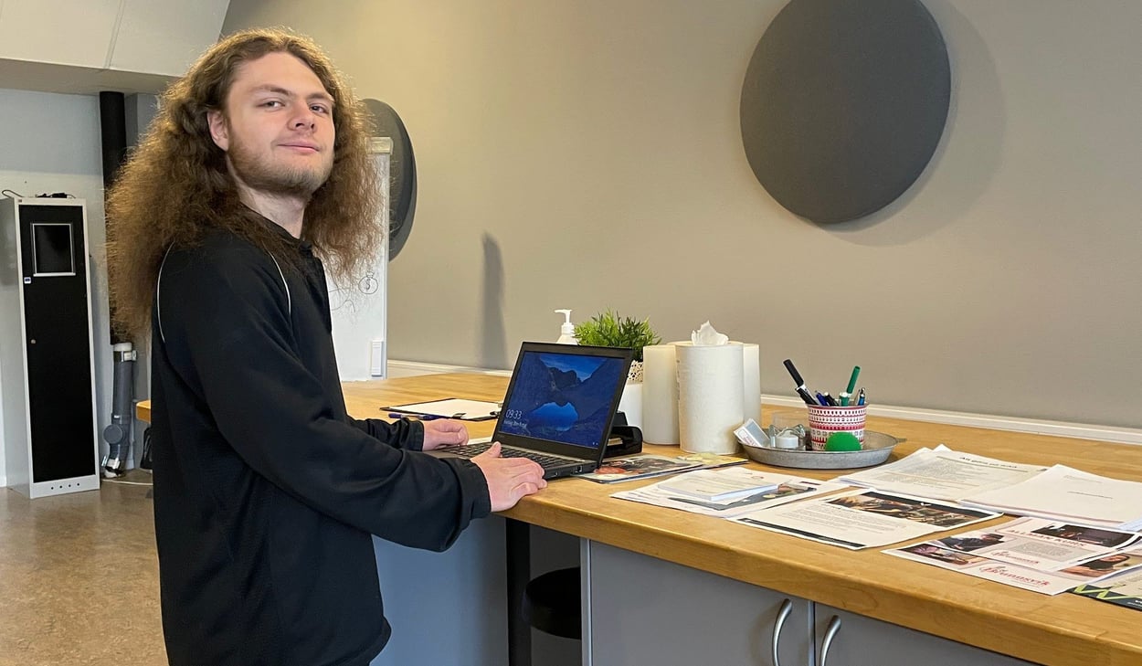 Person vid en dator ståendes vid en arbetsbänk med papper liggandes på sidan.