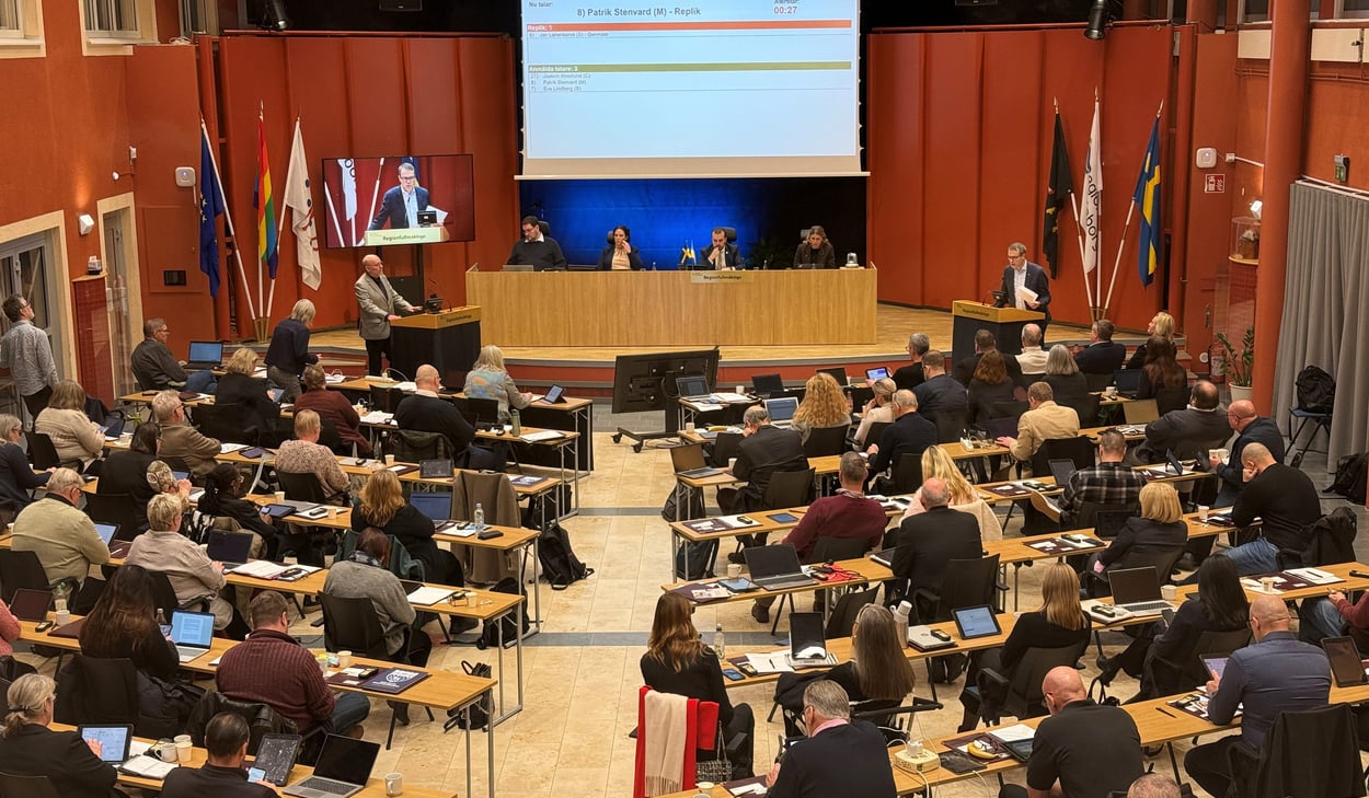 En fullsatt regionfullmäktigesal med deltagare vid bärbara datorer och dokument på borden. En person talar vid en talarstol, och en presentation visas på en stor skärm med text och en talartidräknare. Jan Lahenkorva och Patrik Stenvard debatterar.