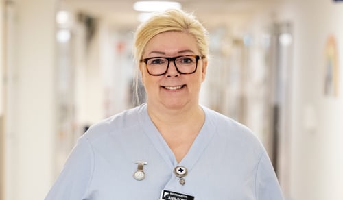 En kvinna i vårdkläder och glasögon står i en korridor.