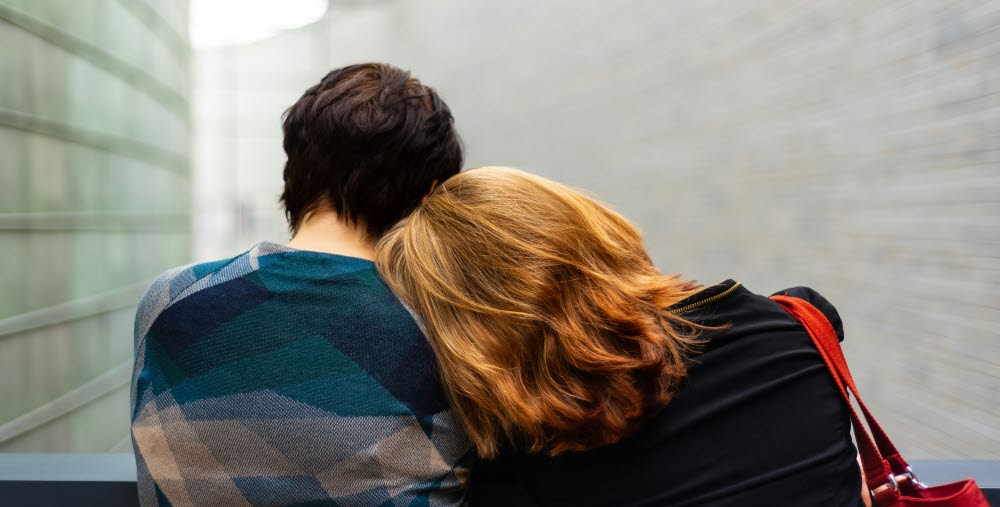 Två personer sitter med ryggen mot kameran; den ena vilar sitt huvud mot den andras axel.
