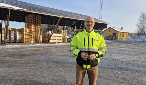 Mattias Ringström iklädd gul varseljacka står på en industriell gårdsplan med staplade träpallar och ett skjul i bakgrunden.