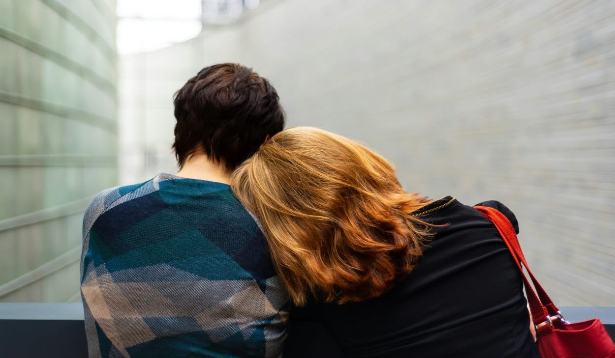 Två personer sitter med ryggen mot kameran; den ena vilar sitt huvud mot den andras axel.