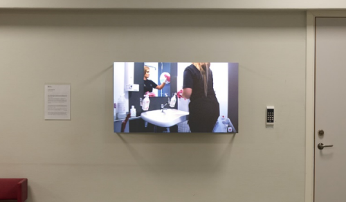 Skärm visar en videoinstallation med två personer vid ett handfat, uppsatt på en vägg i en korridor med bänk och stängd dörr.