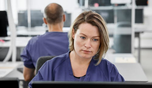 Serious female doctor using computer. Young medical professional sitting at desk in control room. She is in scrubs at hospital.