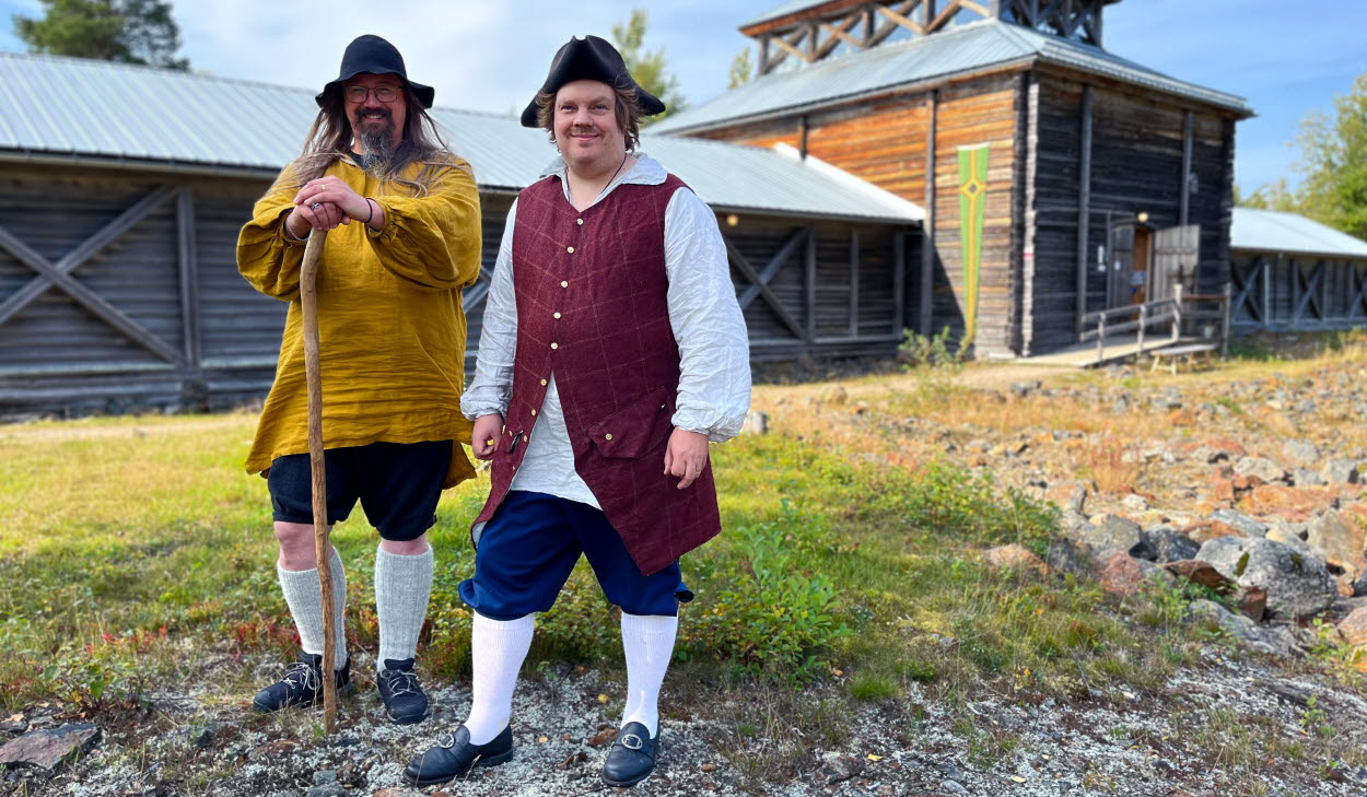 Två män i helfigur klädda i 1700-talskläder står på stenig mark framför en träbyggnad i solsken.