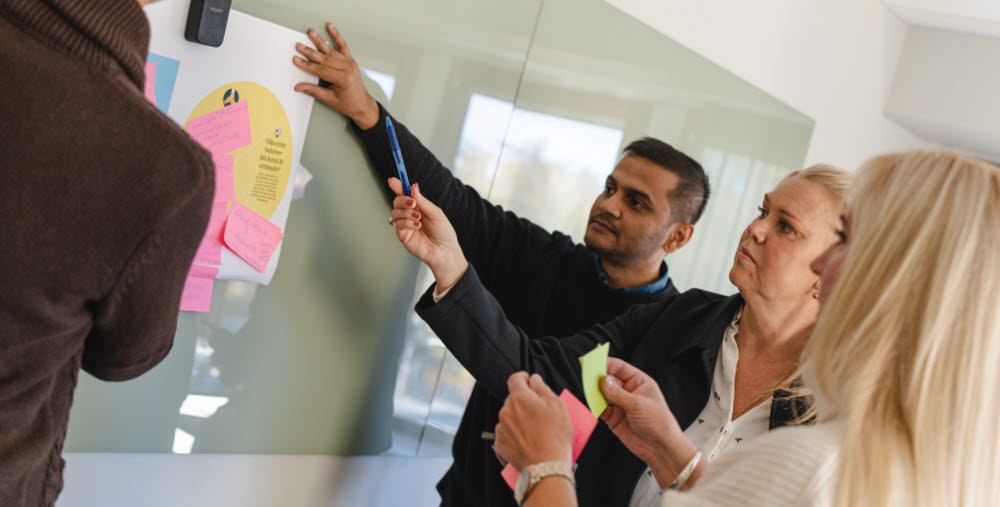 Tre personer framför en whiteboard diskuterar och sätter upp post-it lappar