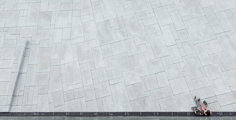 Två personer sitter på marken intill en speglande vägg på ett stort ljusgrått stenlagt torg.