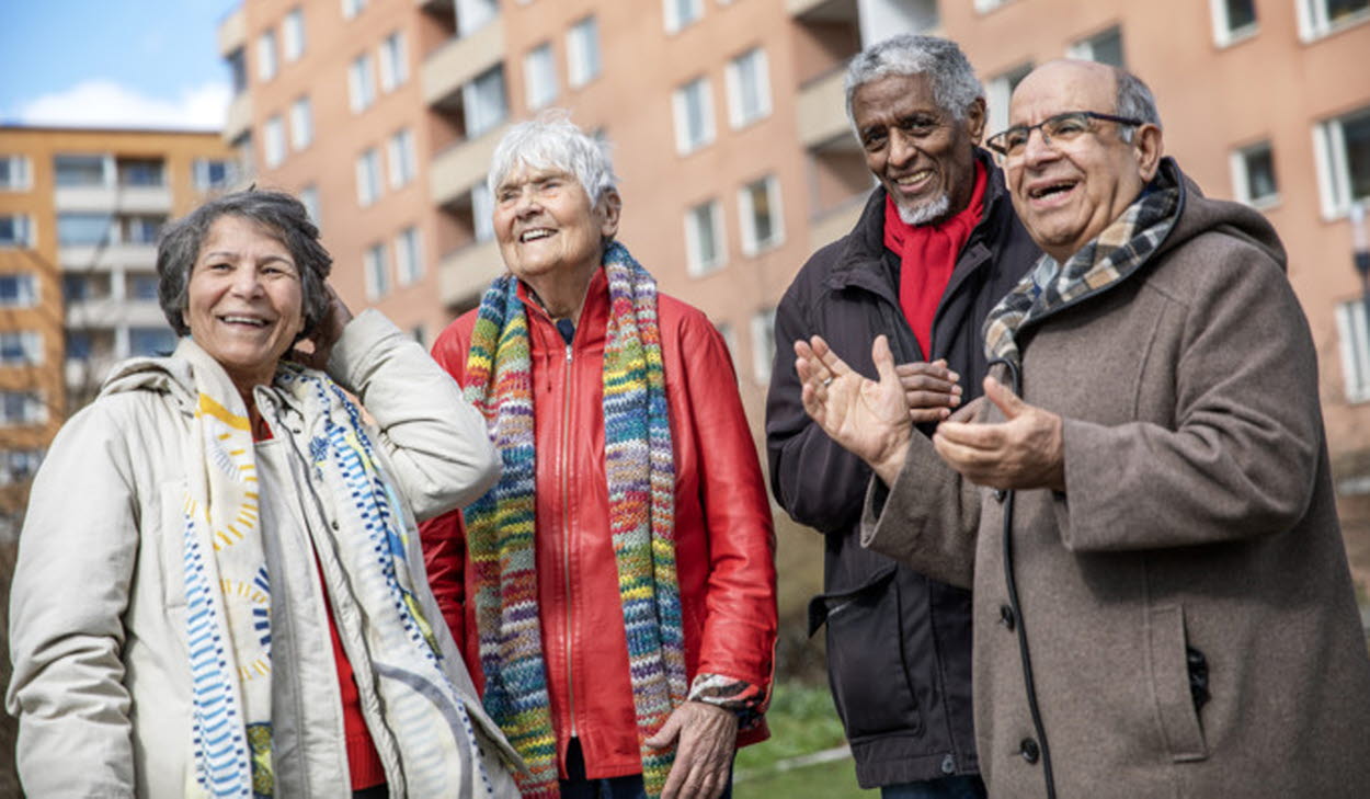 Fyra äldre personer står utomhus och samtalar framför ett bostadshus.